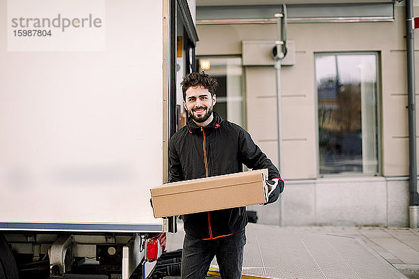 Porträt eines selbstbewussten jungen Zustellers mit einer Kiste  die an einen Lastwagen gelehnt ist