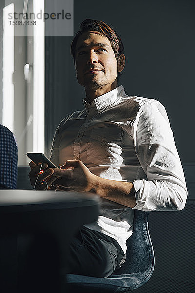 Selbstbewusster Geschäftsmann sitzt mit Smartphone am Konferenztisch im Sitzungssaal