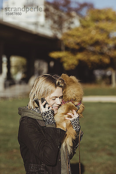 Lächelnde blonde Frau telefoniert mit einem Mobiltelefon  während sie an einem sonnigen Tag Pommern im Park trägt