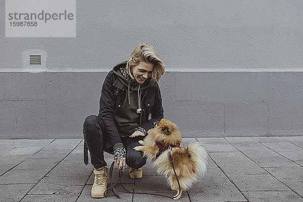 Lächelnde blonde Frau kauert  während sie mit dem pommerschen Hund an der Wand spielt