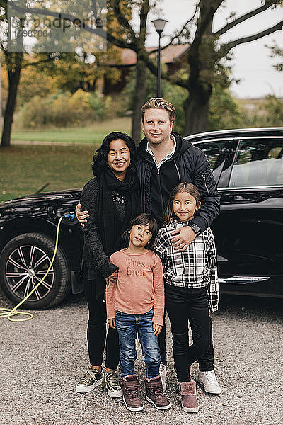 Porträt von Vater und Mutter mit Töchtern vor Elektroauto stehend