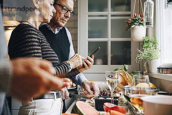 Ältere Frau zeigt einem Mann ein Smartphone  während sie in der Küche das Abendessen zubereitet