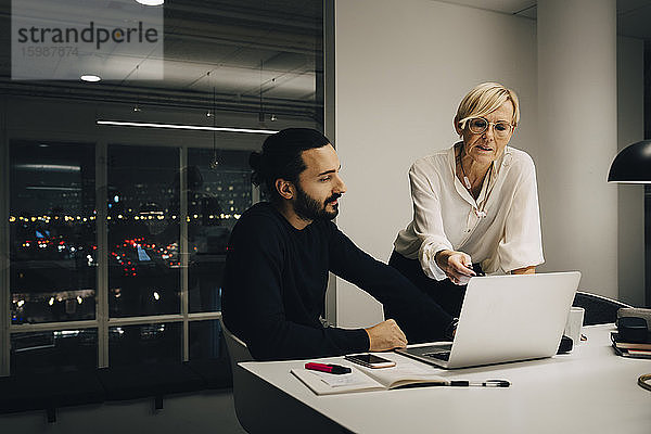 Männliche und weibliche Fachkräfte planen ihre Strategie  während sie über einen Laptop in einem beleuchteten Büro diskutieren