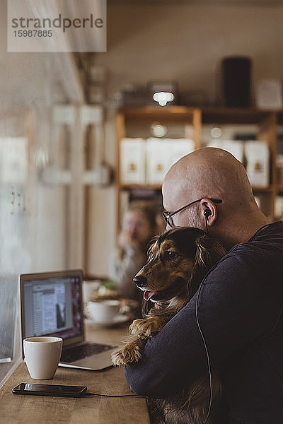 Kahlköpfiger Mann sitzt mit Hund und benutzt Laptop im Café