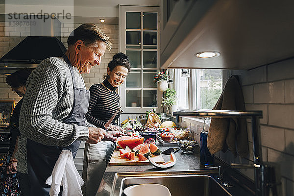 Lächelnde ältere männliche und weibliche Freunde schneiden Früchte an der Küchentheke