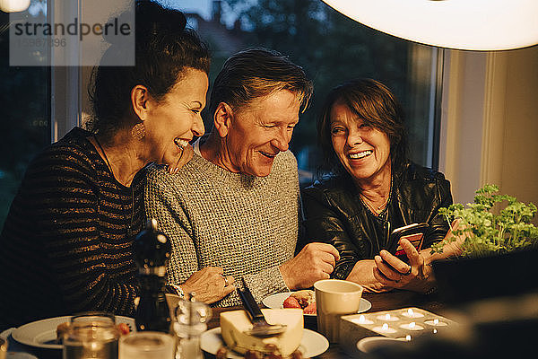 Lächelnde ältere Frau teilt ihr Smartphone mit Freunden  während sie während der Dinnerparty am Esstisch sitzt