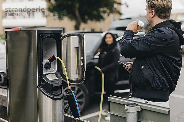 Mann trinkt Kaffee  während er an einer Ladestation für Elektroautos steht