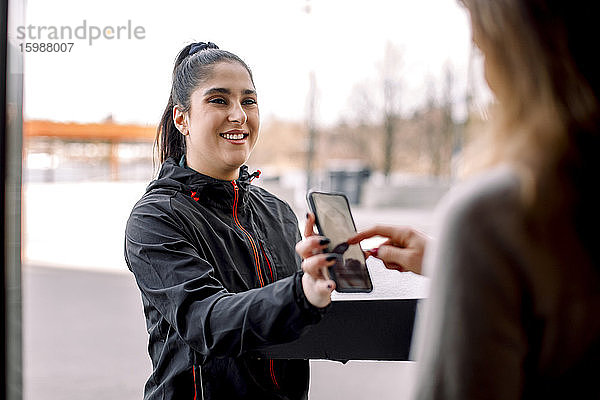 Frau unterschreibt auf Smartphone  während sie ein Paket von einer Zustellerin erhält