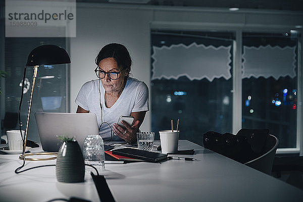 Selbstbewusste reife Unternehmerin nutzt Laptop  während sie im Büro mit Smartphone sitzt