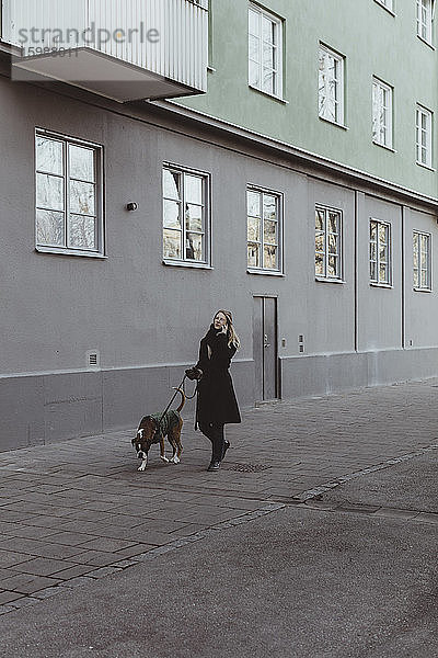 Junge Frau in voller Länge geht mit Hund auf einem Fußweg durch ein Gebäude in der Stadt