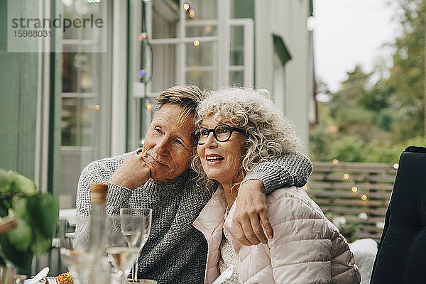 Nachdenklicher älterer Mann sitzt mit dem Arm um die Frau am Esstisch während der Gartenparty