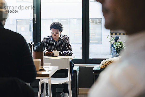 Junger Kunde benutzt Mobiltelefon  während er mit Kaffeetasse am Tisch im Cafe sitzt