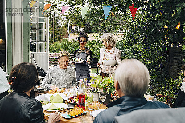 Aktive ältere Frauen bringen Freunden  die während einer Gartenparty am Esstisch sitzen  das Essen