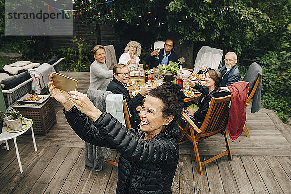 Lächelnde ältere Frau  die während der Dinnerparty mit Freunden am Esstisch im Hinterhof sitzt