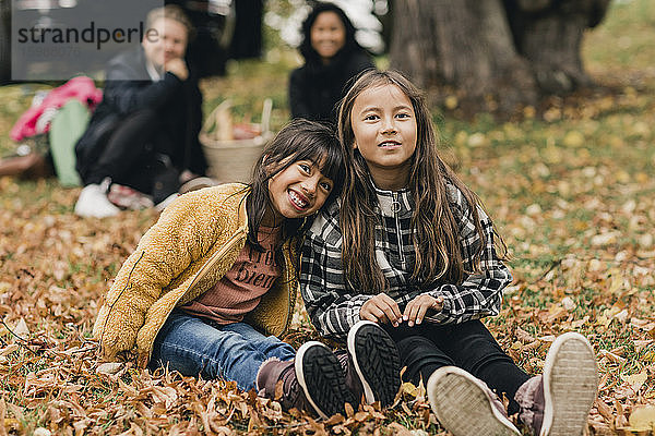 Porträt von lächelnden Schwestern  die während des Picknicks mit den Eltern auf dem Herbstlaub sitzen