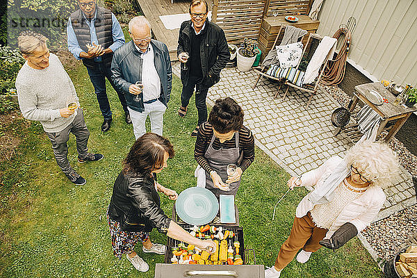 Schrägaufnahme von lächelnden älteren Männern  die während der Gartenparty auf Frauen blicken  die das Abendessen auf dem Grill zubereiten