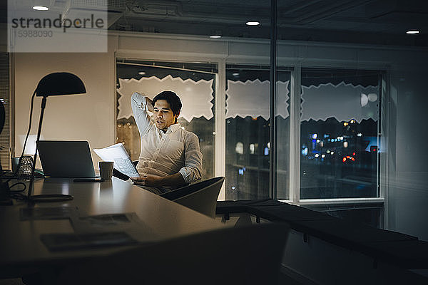 Selbstbewusster männlicher Unternehmer liest Dokument  während er nachts in einem dunklen Büro sitzt