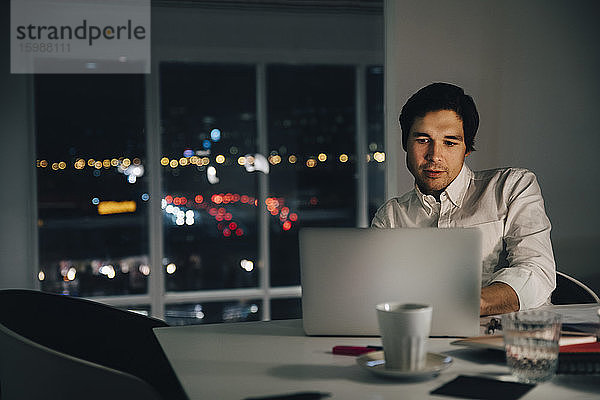 Selbstbewusster Geschäftsmann mit Laptop  während er am Schreibtisch an einem kreativen Arbeitsplatz sitzt