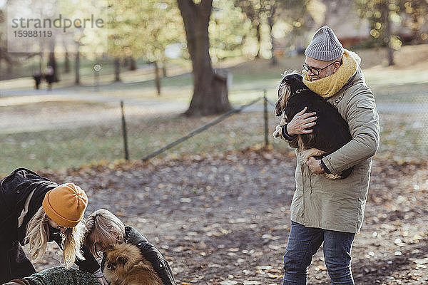 Lächelnder Mann trägt Hund  während er im Park auf Frauen und Haustiere zugeht