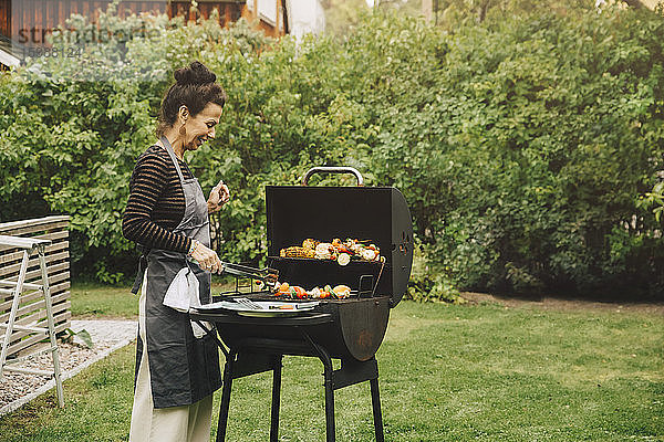 Seitenansicht einer lächelnden Frau beim Kochen des Abendessens auf dem Grill im Hinterhof während einer Gartenparty
