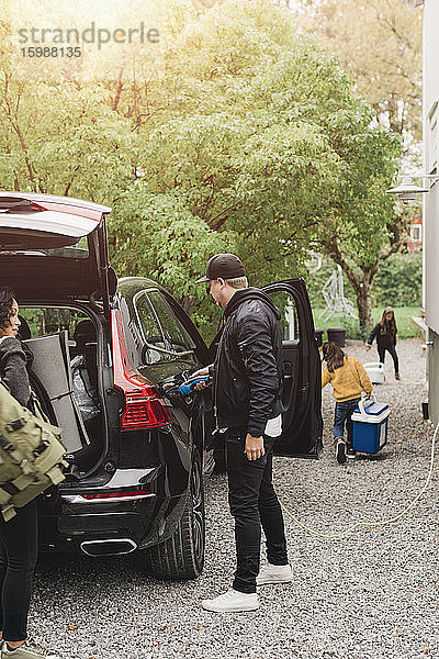 Mittelgroßer erwachsener Mann lädt Elektroauto auf  während die Familie beim Picknick Gepäck einlädt