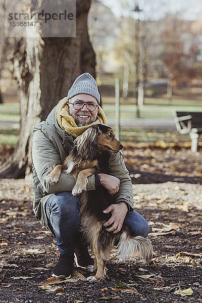 Porträt eines lächelnden Mannes  der einen Hund umarmt  während er im Herbst im Park kauert