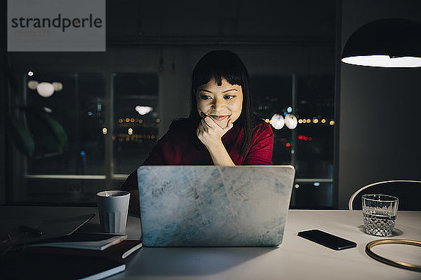 Zuversichtliche Geschäftsfrau schaut auf den Laptop  während sie mit der Hand am Kinn sitzend bis spät im Büro arbeitet