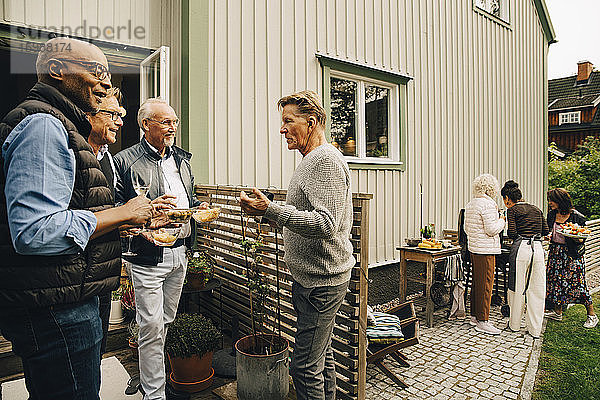 Ältere Männer unterhalten sich und genießen Snacks  während Frauen im Hinterhof das Abendessen vorbereiten