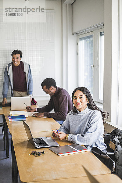 Lächelnde junge Frau schaut weg  während sie am Schreibtisch im Klassenzimmer sitzt