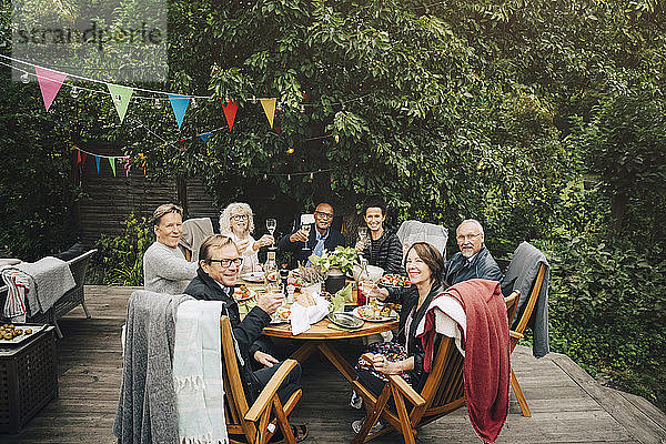 Porträt lächelnder aktiver älterer Männer und Frauen mit feierlichem Toast  die während der Party am Esstisch im Hinterhof sitzen