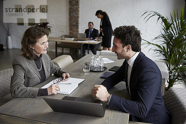 Zuversichtliche ältere Anwältin lächelt  während sie während eines Treffens im Büro mit einem jungen Geschäftsmann über einen Laptop diskutiert