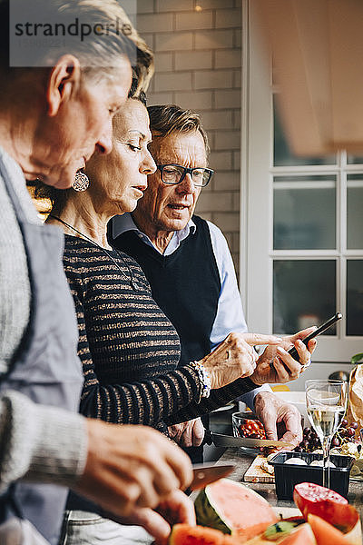 Ältere Frau diskutiert mit männlichen Freunden über Smartphone  während sie zu Hause in der Küche steht