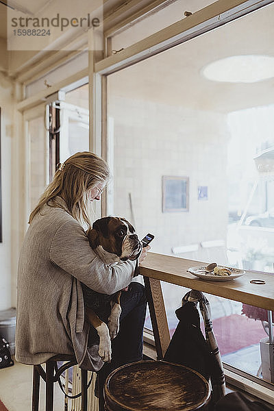 Seitenansicht einer jungen Tierhalterin  die ein Smartphone benutzt  während sie mit einem Boxerhund im Café sitzt