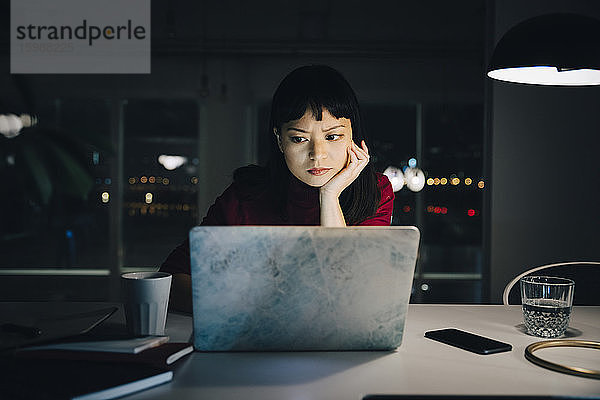 Engagierte junge Geschäftsfrau mit einer Hand am Kinn  die einen Laptop benutzt  während sie spät im Büro arbeitet