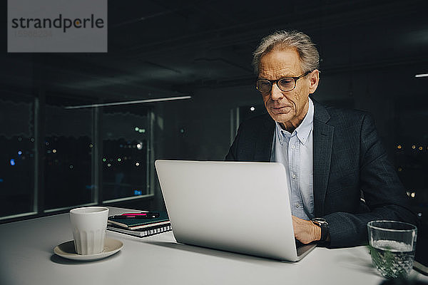 Selbstbewusster Senior-Geschäftsmann mit Laptop am beleuchteten Schreibtisch im Kreativbüro
