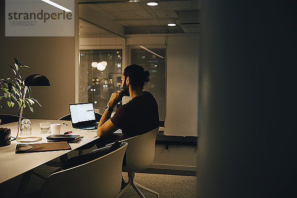 Seitenansicht eines engagierten männlichen Mitarbeiters  der spät am beleuchteten Schreibtisch im Coworking Space arbeitet