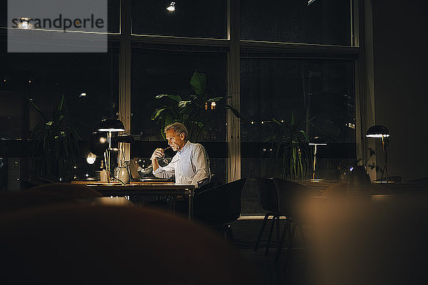 Nachdenklicher Geschäftsmann  der spät arbeitet  während er mit seinem Laptop an einem dunklen Arbeitsplatz sitzt