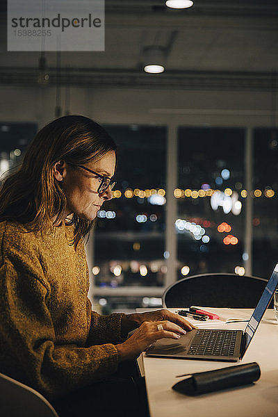 Seitenansicht eines weiblichen Mitarbeiters  der spät arbeitet  während er seinen Laptop am beleuchteten Schreibtisch im Büro benutzt