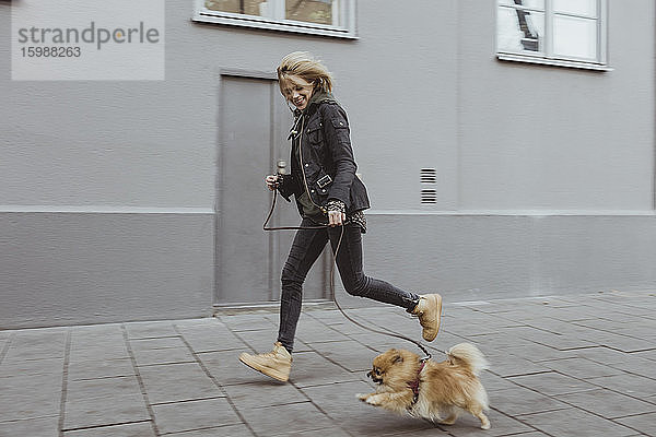Seitenansicht in voller Länge einer glücklichen Frau  die mit ihrem pommerschen Hund auf einem Fußweg durch ein Gebäude in der Stadt läuft