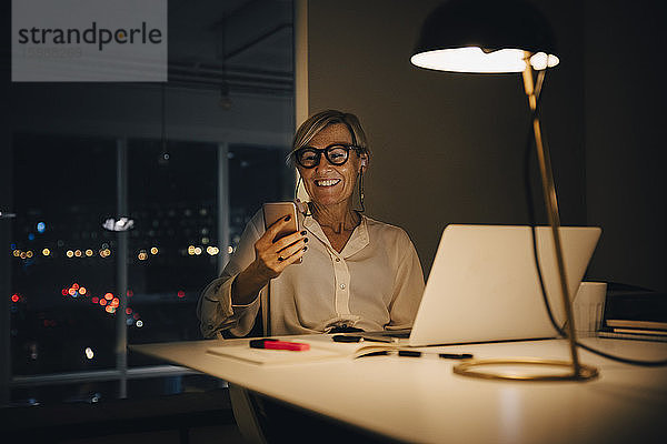 Lächelnde Geschäftsfrau mit Smartphone  während sie mit Laptop am beleuchteten Schreibtisch im Coworking Space sitzt