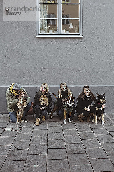 Porträt in voller Länge von lächelnden Haustierbesitzern mit Hunden auf einem Fußweg gegen eine Mauer in der Stadt