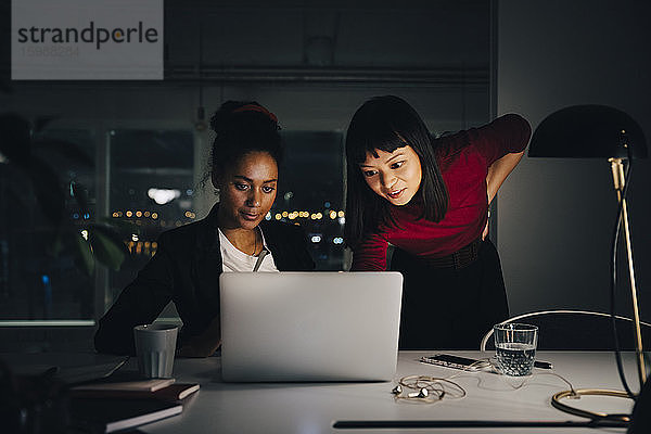 Zuversichtliche weibliche Geschäftskollegen diskutieren am Laptop  während sie spät im Kreativbüro arbeiten