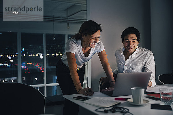 Lächelnde männliche und weibliche Kollegen diskutieren am Laptop  während sie spät im Kreativbüro arbeiten