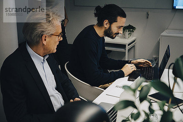 Leitender Geschäftsmann schaut während einer Sitzung am Konferenztisch im Sitzungssaal zu  während er von einem männlichen Kollegen am Laptop sitzt