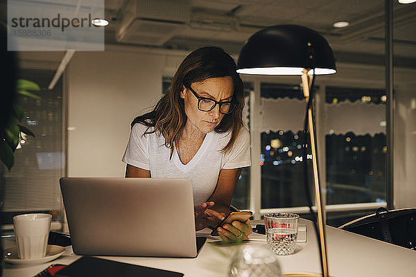 Selbstbewusste Unternehmerin  die ein Smartphone benutzt  während sie nachts mit ihrem Laptop an einem beleuchteten Schreibtisch am Arbeitsplatz sitzt