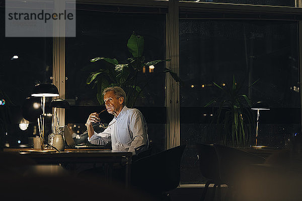 Nachdenklicher älterer männlicher Fachmann schaut weg  während er in einem dunklen Büro am Schreibtisch sitzt