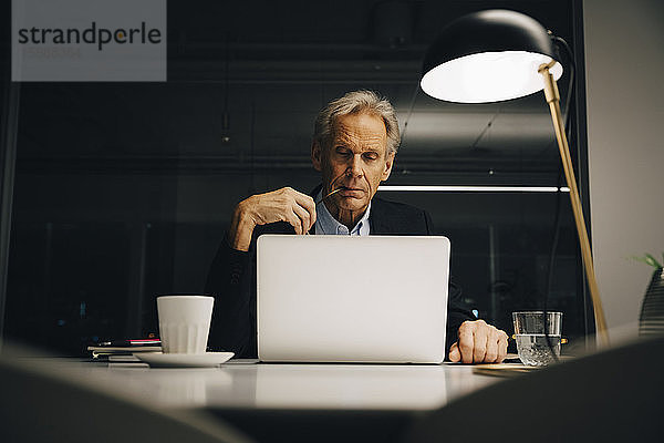 Engagierter älterer männlicher Unternehmer schaut auf den Laptop  während er bis spät in der kreativen Arbeitswelt arbeitet