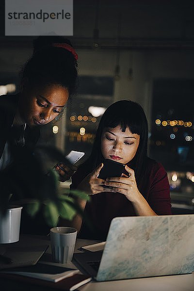 Zuversichtliche Kolleginnen  die Smartphones benutzen  während sie spät am Coworking Space arbeiten