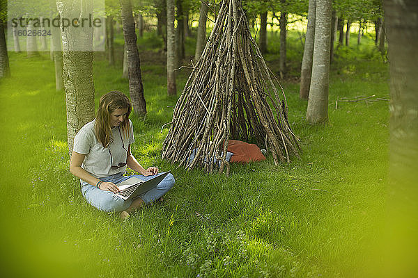 Junge Frau mit Laptop am Ast-Tipi im Wald