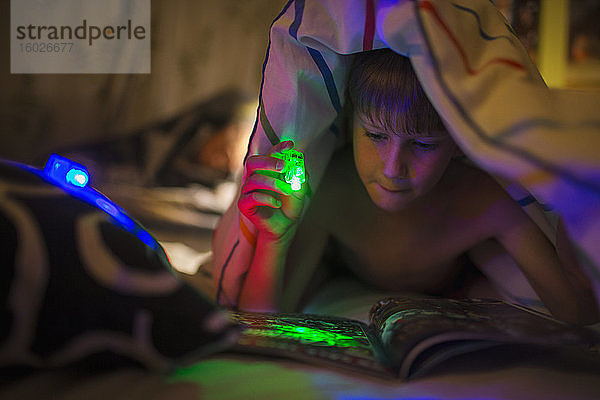 Junge mit grüner Taschenlampe liest Zeitschrift unter einer Decke